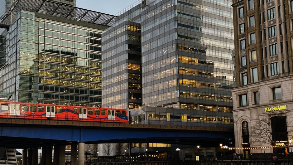 A city scene featuring a red and blue urban train crossing a blue railway bridge set against modern glass office buildings with reflective surfaces, illuminated windows, and sleek architectural design