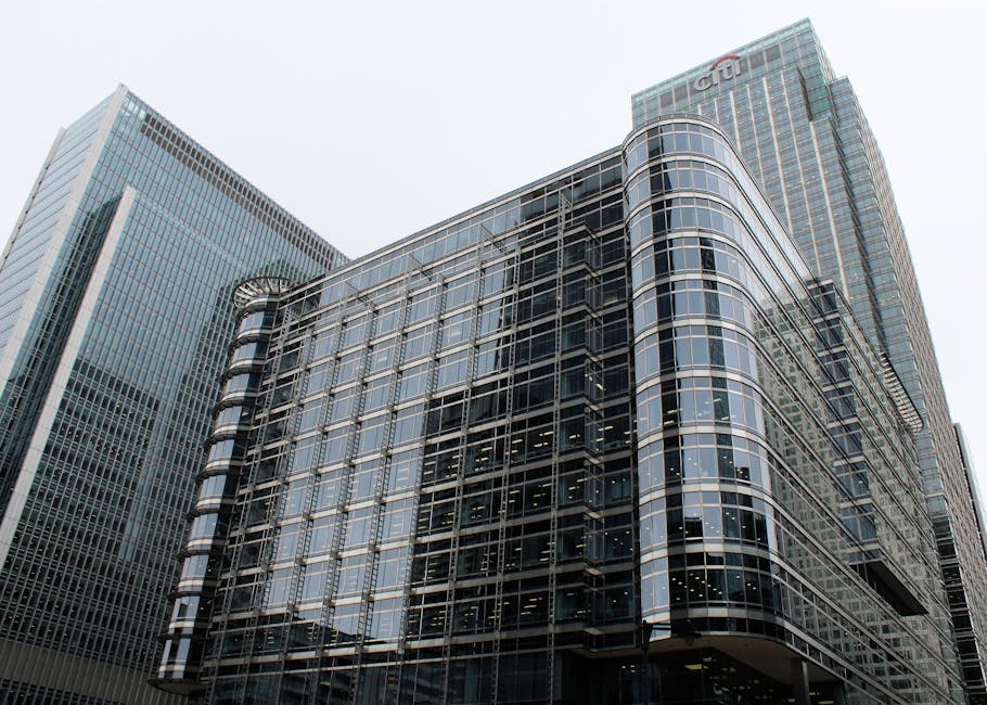 A city scene featuring a red and blue urban train crossing a blue railway bridge set against modern glass office buildings with reflective surfaces, illuminated windows, and sleek architectural design