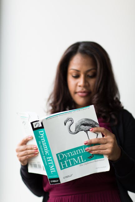 A woman with medium skin tone and dark, wavy hair is holding a book titled 'Dynamic HTML' with a picture of a flamingo on the cover. She is dressed in a black blazer over a maroon top and stands again