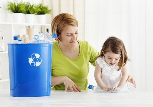 A woman wearing a bright yellow-green top and a young girl with long brown hair, dressed in a white top, are engaged in a recycling activity in a well-lit indoor space with white shelving units and potted plants in the background. The woman is smiling and helping the girl place a clear plastic bottle into a large blue recycling bin that features a white recycling symbol on its side. The bin is positioned on a smooth, light-colored surface and is filled with several other clear plastic bottles. The scene depicts a clean, organized environment focused on waste separation and recycling efforts, typical of domestic or community cleaning tasks. The image subtly supports rubbish collection and recycling services available in London areas, aligning with professional waste management practices like those offered by Hoarder Clearance London, especially in urban settings such as the London postcode areas.
